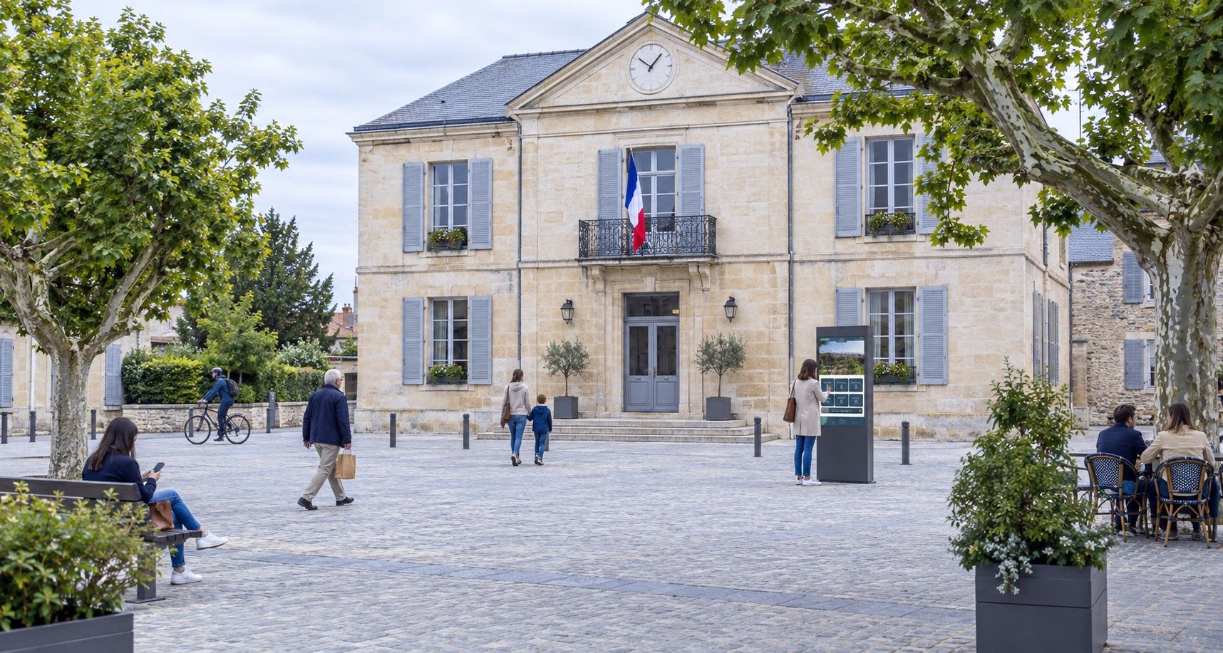 A busy village square in front of a town hall, with a municipal information screen among the other elements of the square